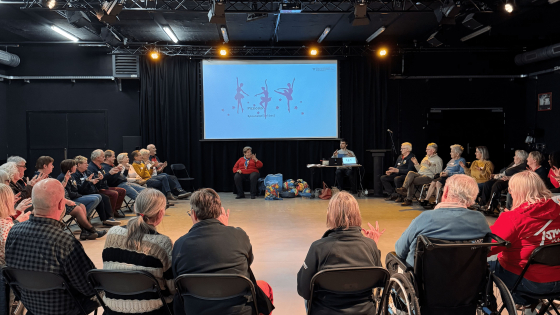 Een groep mensen in een kring kijkt naar een presentatie op groot scherm